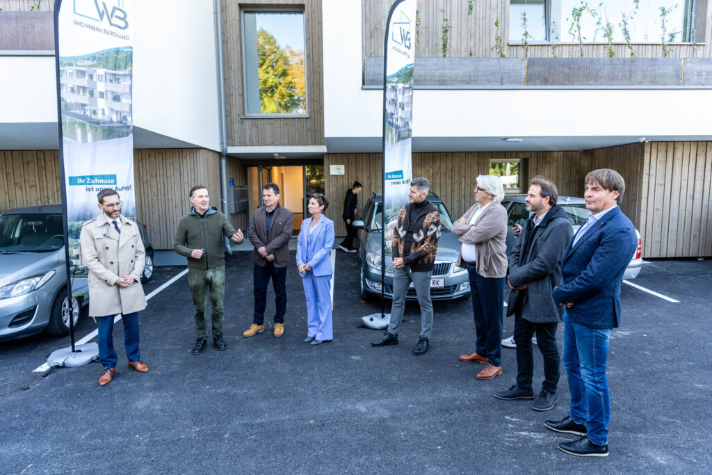 Gäste der Wohnungsübergabe vor dem Eingang des neuen Wohnhauses in Grödig, daneben Roll-ups der Wohnbau Bergland